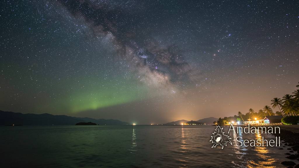 A wide view of the dark night sky above a calm tropical coastline at midnight, countless stars and the Milky Way arching overhead in a luminous band, a faint green airglow near the horizon, gentle warm light from a distant fishing village, calm ocean reflecting starlight with gentle ripples